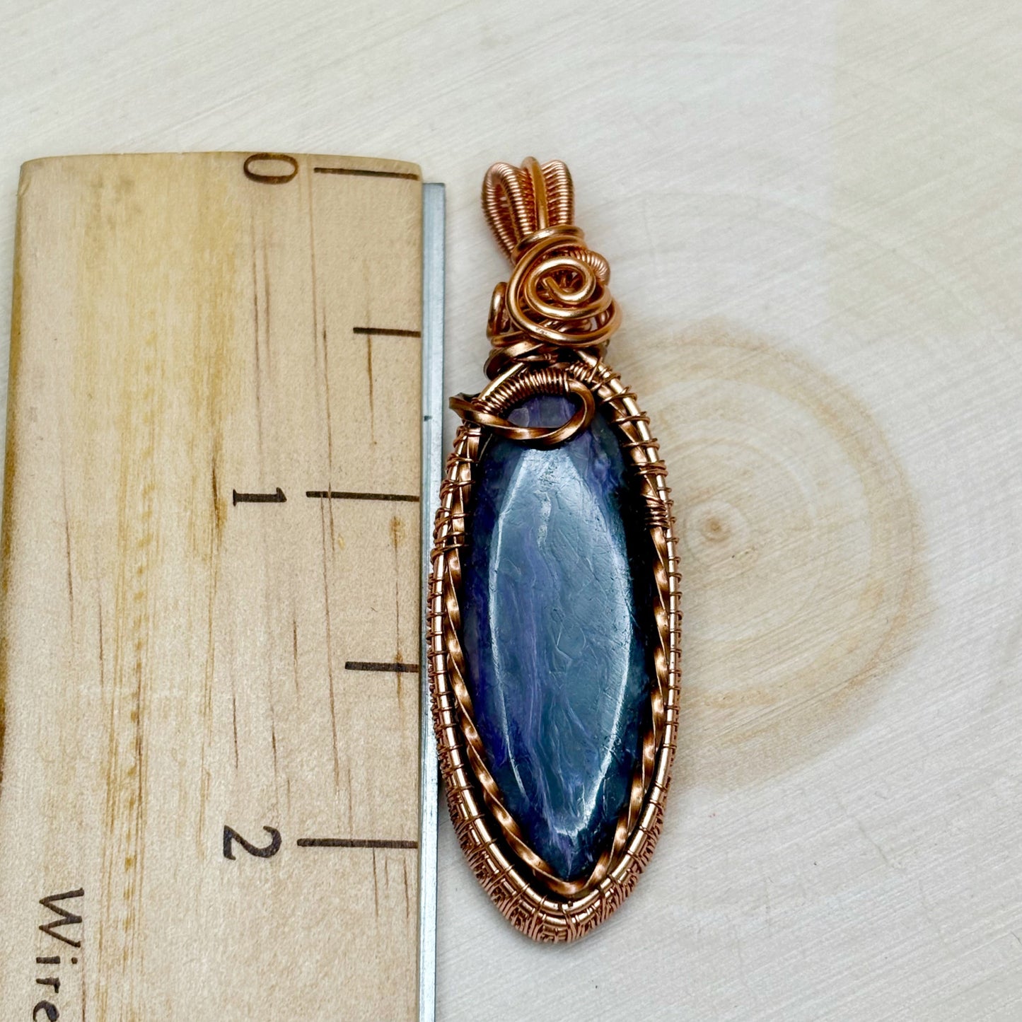 purple pendant with copper wire wrapping on a wooden block with measurement scale