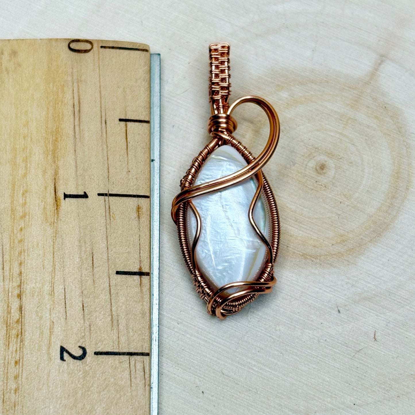 Copper-wrapped pendant with a central mother of pearl shell on a wooden surface with a ruler for scale.