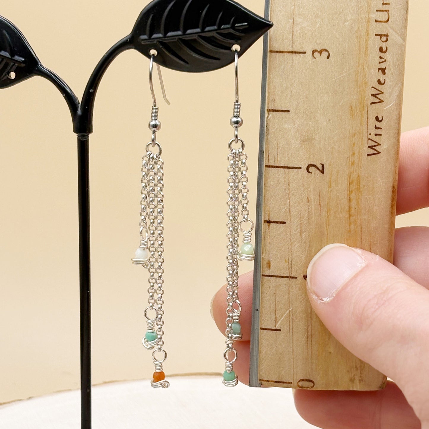 Pair of silver chain earrings with beads held against a wooden ruler for size reference.