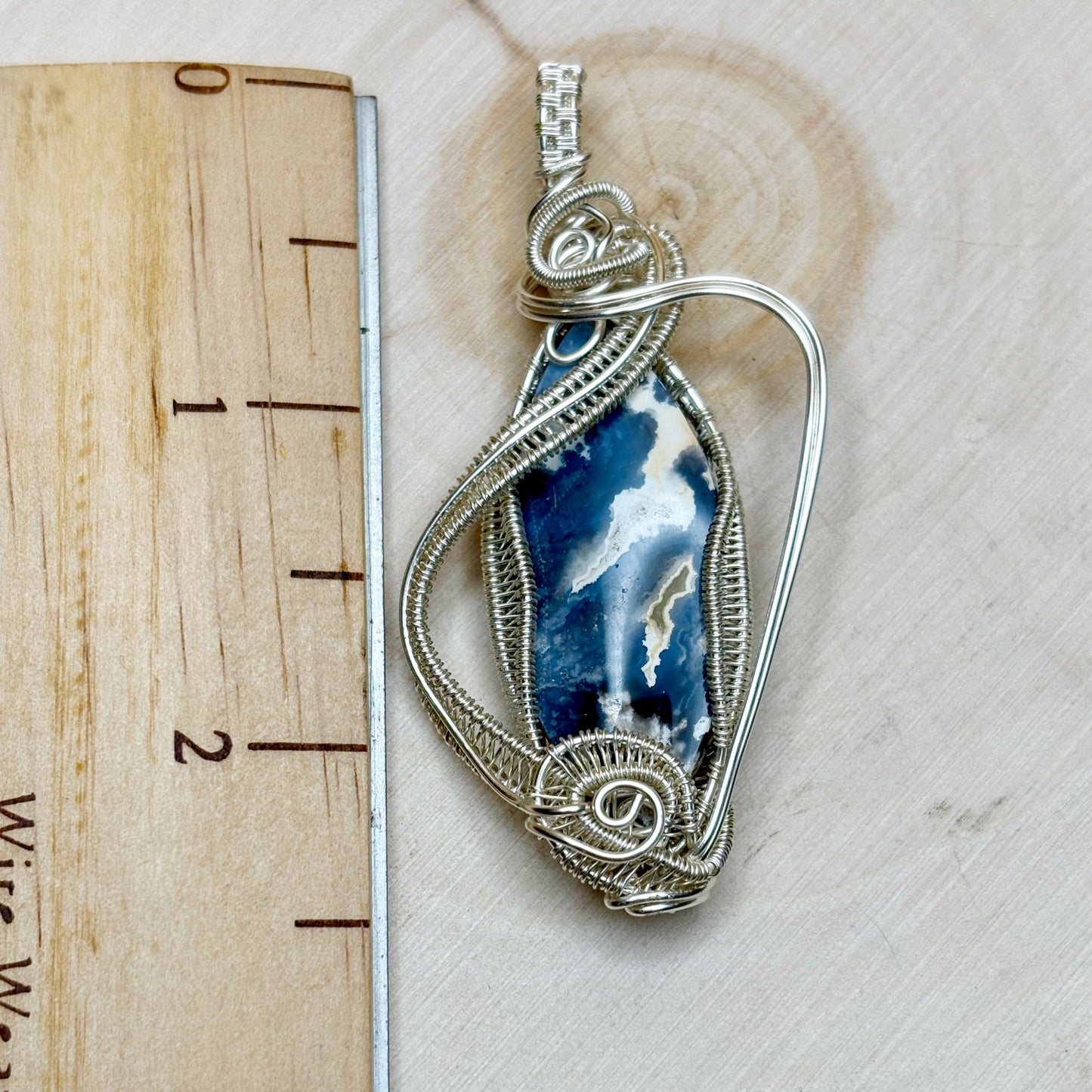 Silver wire-wrapped pendant with a blue and white stone on a wooden surface with a ruler.