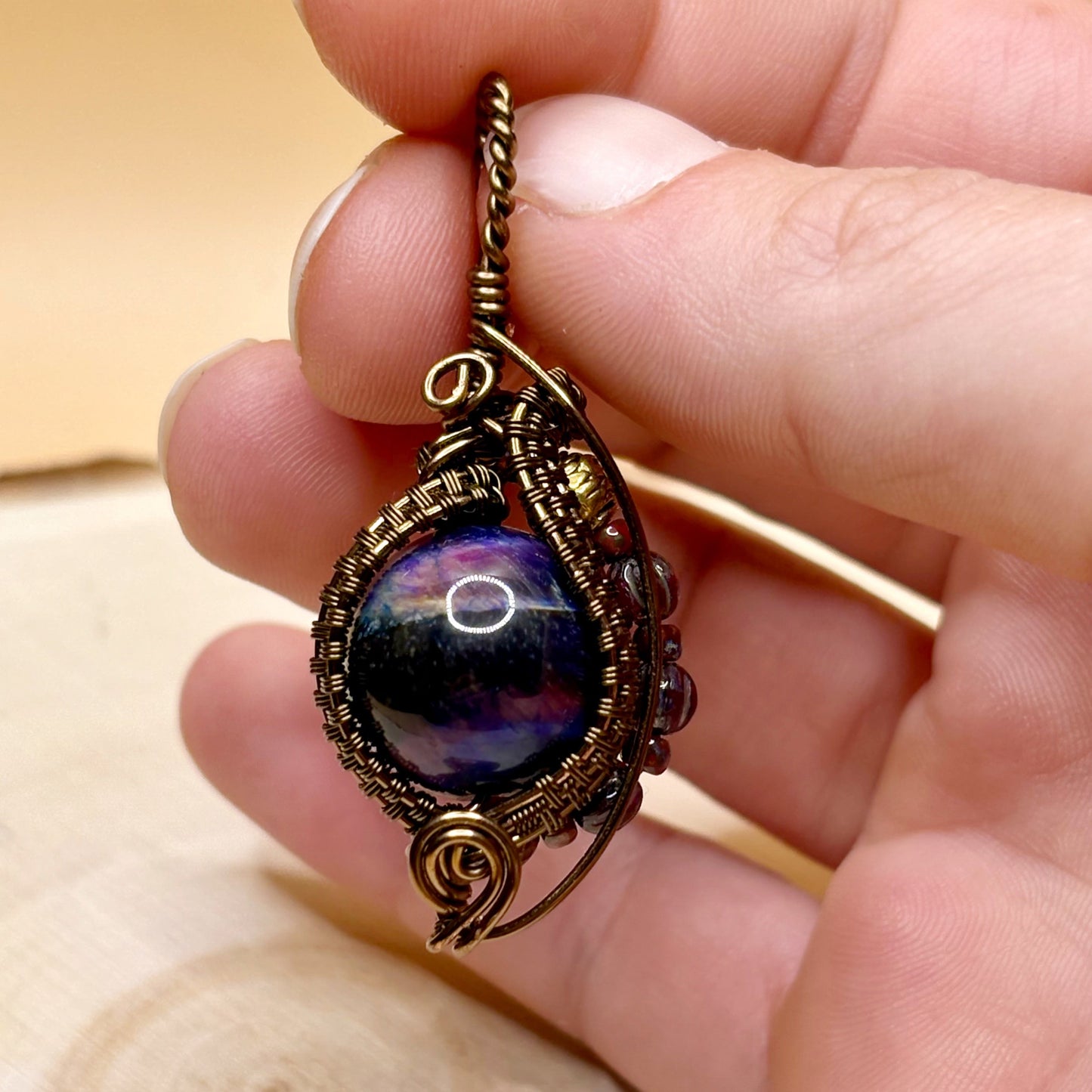 Hand holding a decorative pendant with a stone bead and metal wire wrapping on a wooden surface.
