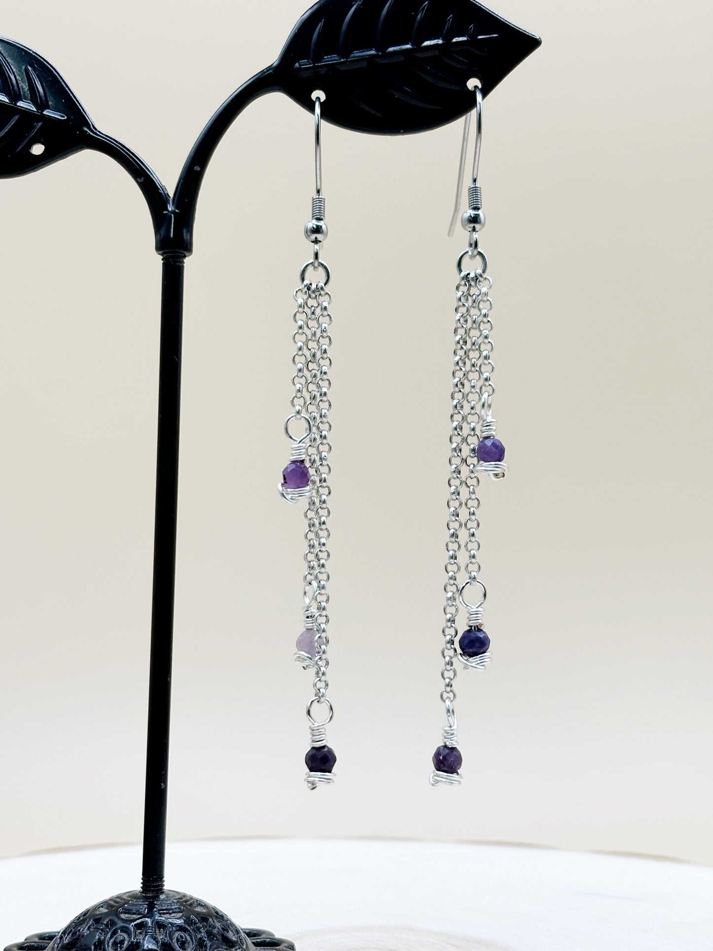 Pair of silver chain earrings with purple beads on a black leaf-shaped stand.