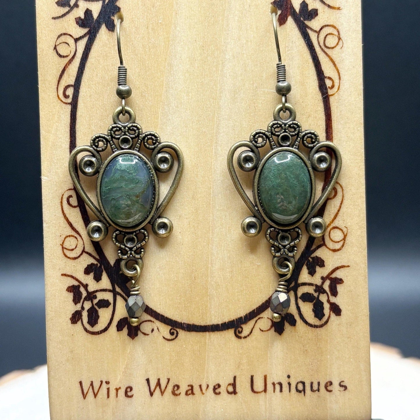Pair of ornate earrings with green stones on a wooden display card with 'Wire Weaved Uniques' branding.