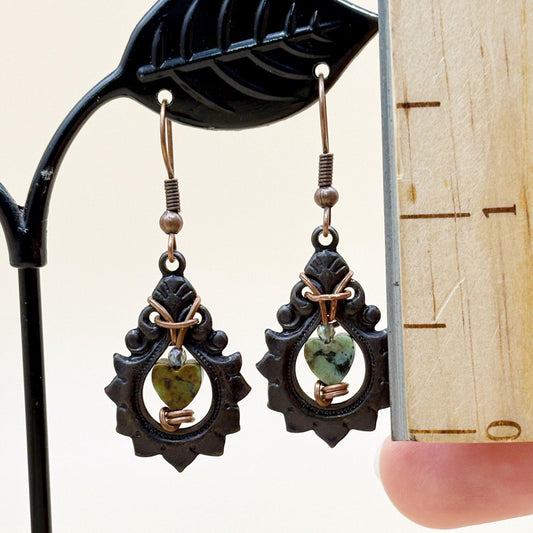 Pair of black-framed earrings with green heart-shaped stones held against a wooden background.