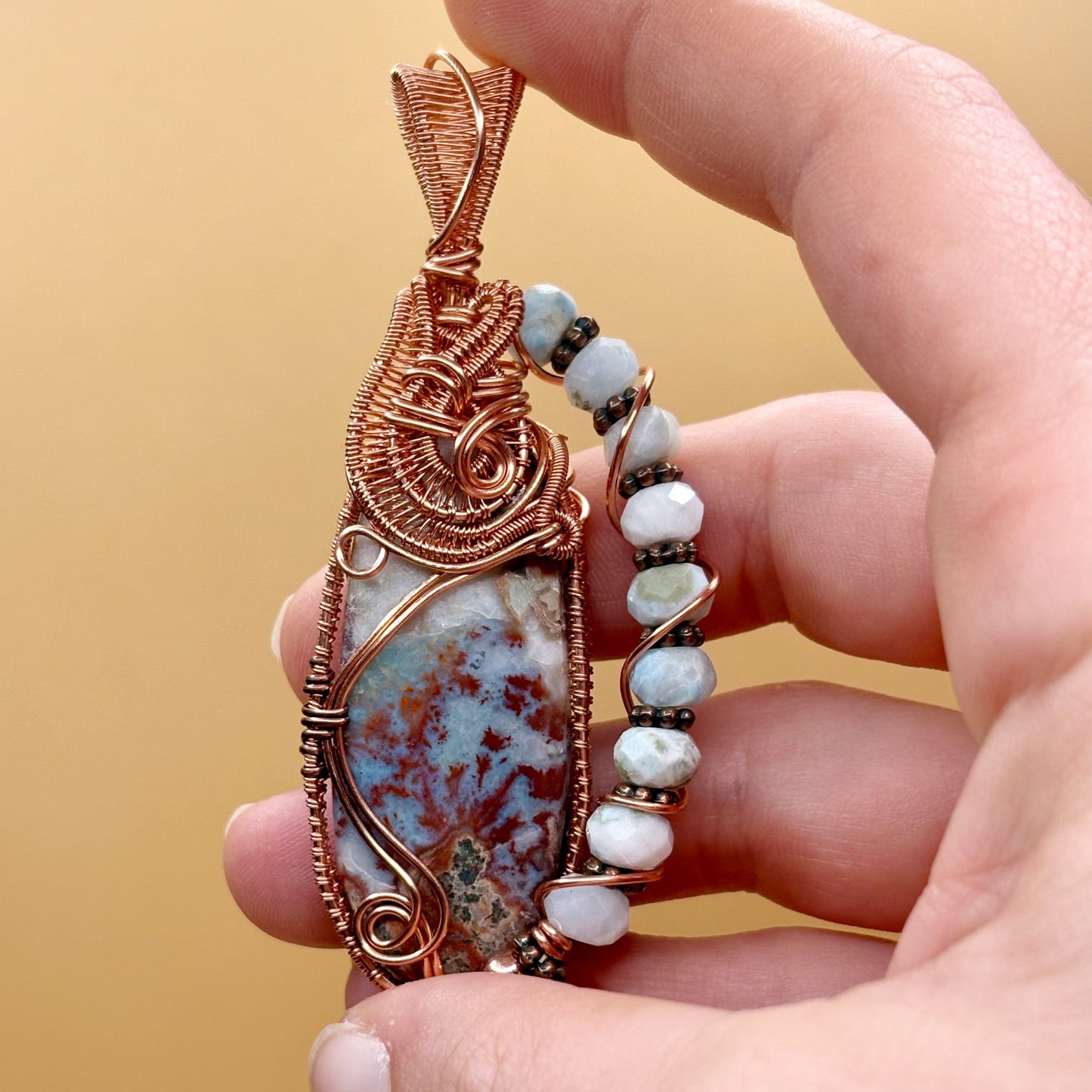 Hand holding a wire-wrapped pendant with larimar stone and blue beads against a beige background