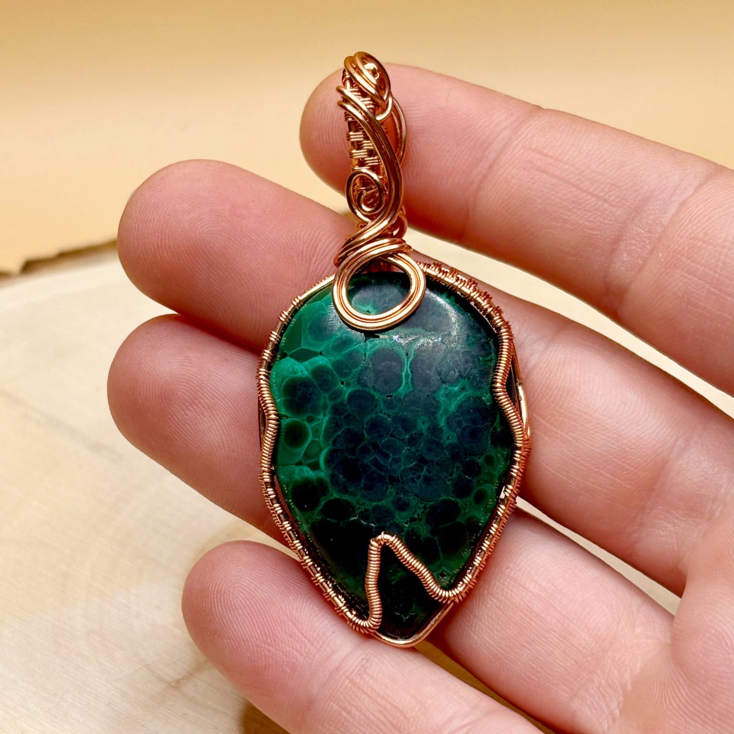 Green stone pendant with copper wire wrapping held in a hand against a beige background