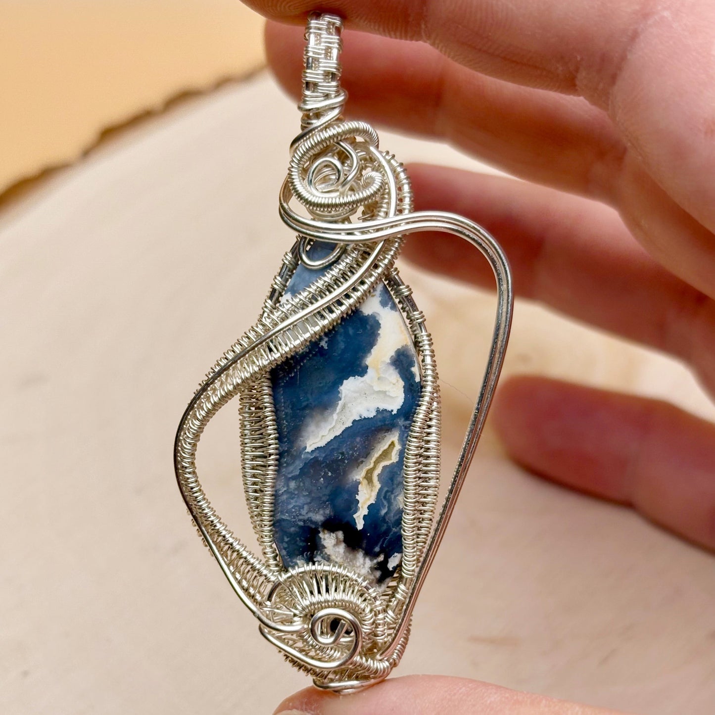 Silver wire-wrapped pendant with a blue and white stone held by a hand against a beige background