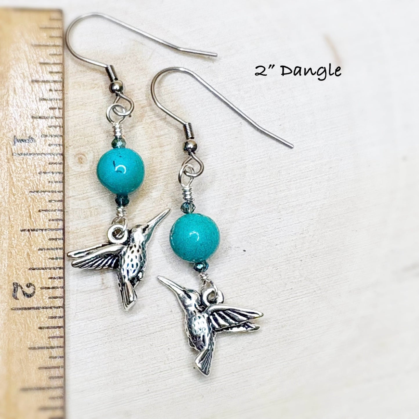 Pair of earrings with turquoise beads and silver hummingbird charms next to a ruler on a white background