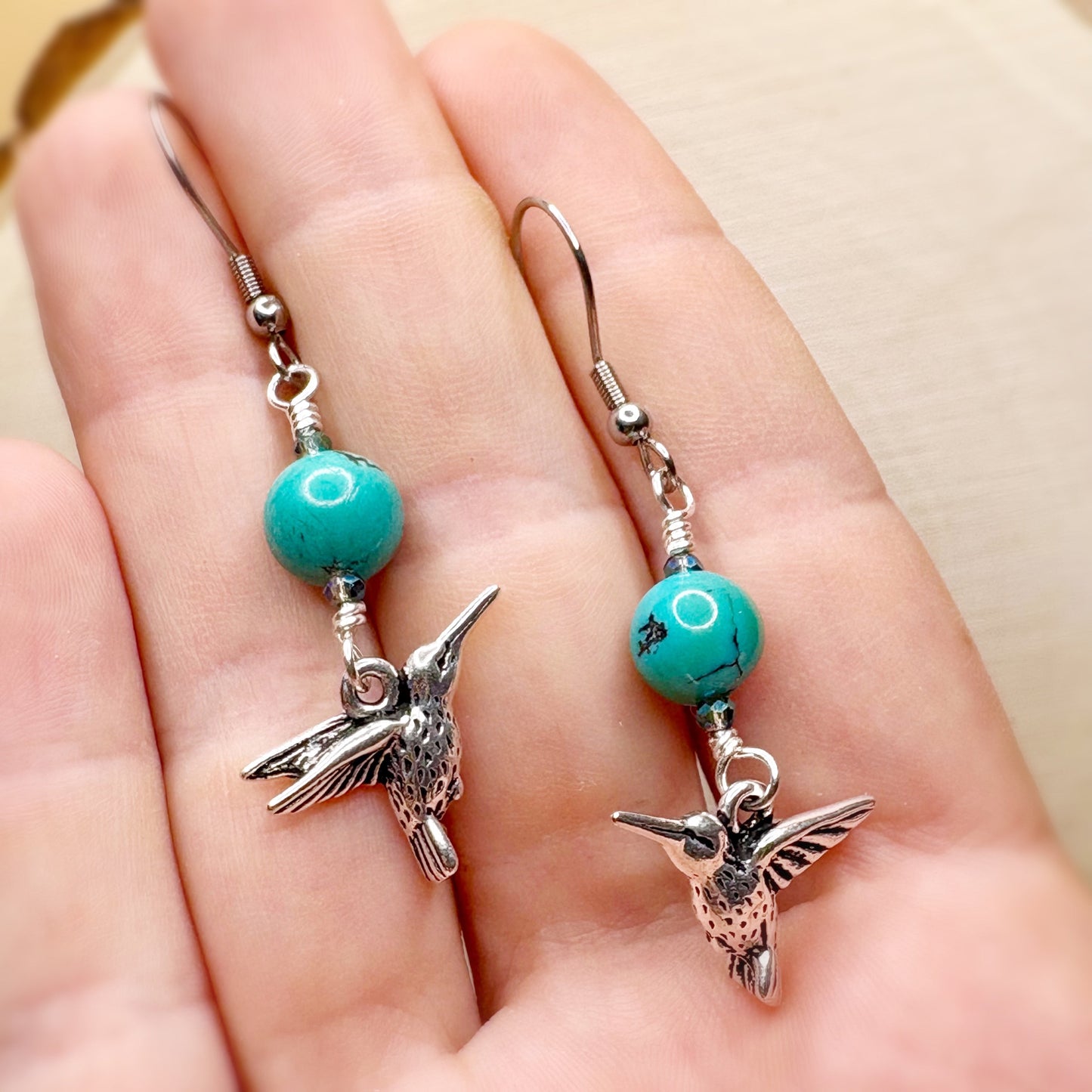 Pair of earrings with turquoise beads and silver hummingbird charms held in a hand.