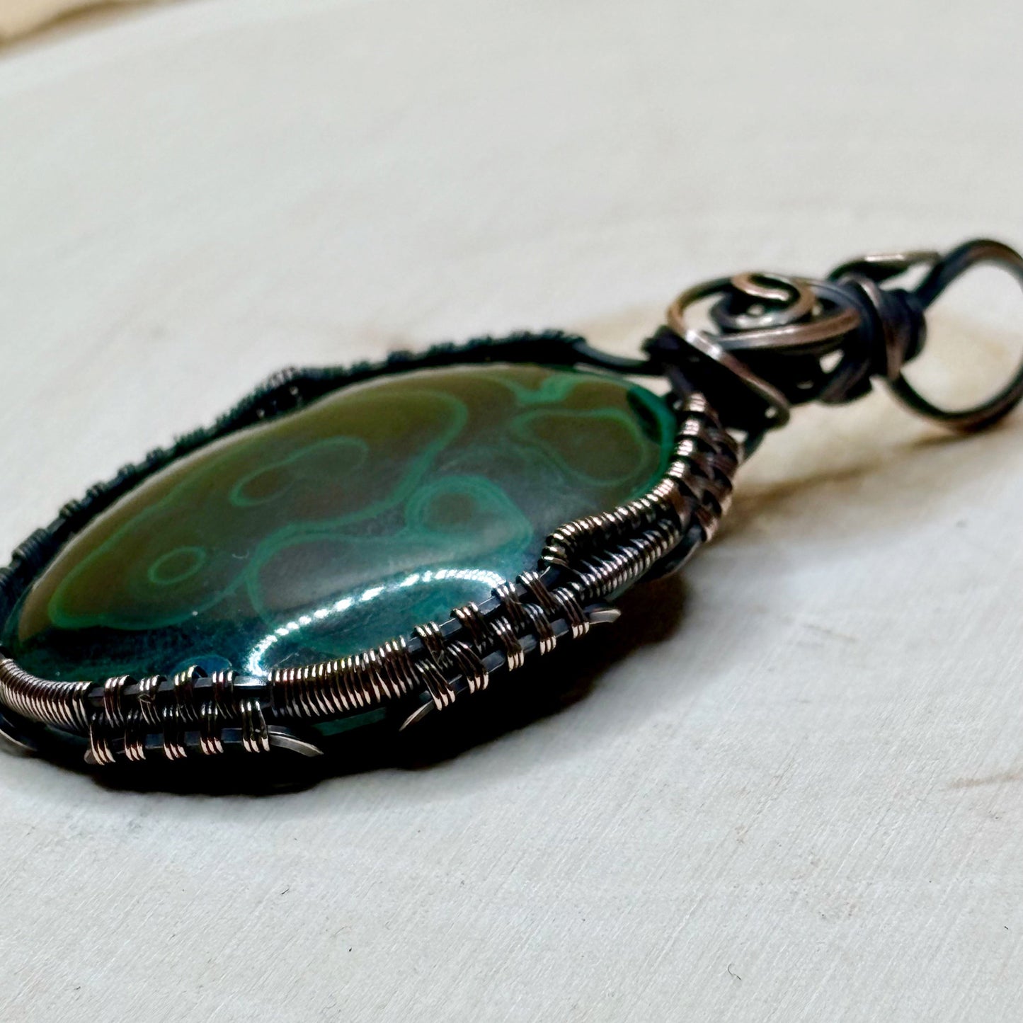 Green stone with some blue wire wrapped in antiqued dark copper with intricate woven frame