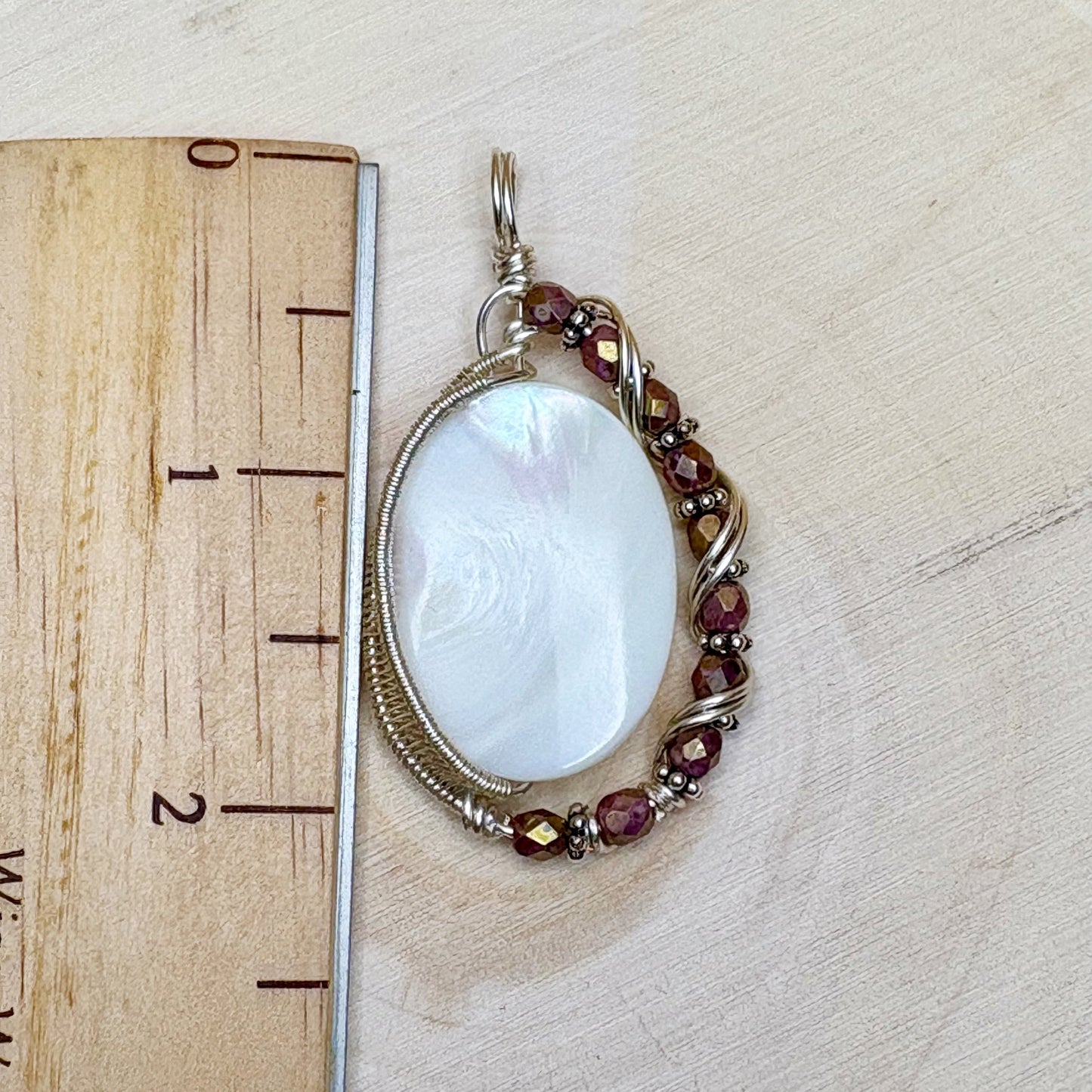 Pendant with a large central mother of pearl and smaller glass beads around it, on a wooden surface with a ruler for scale.