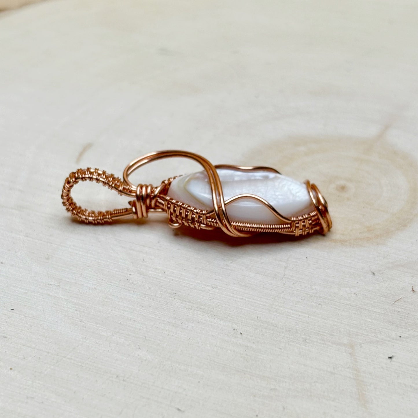 Necklace pendant with a mother of pearl shell wire wrapped in shiny copper on a light wooden background