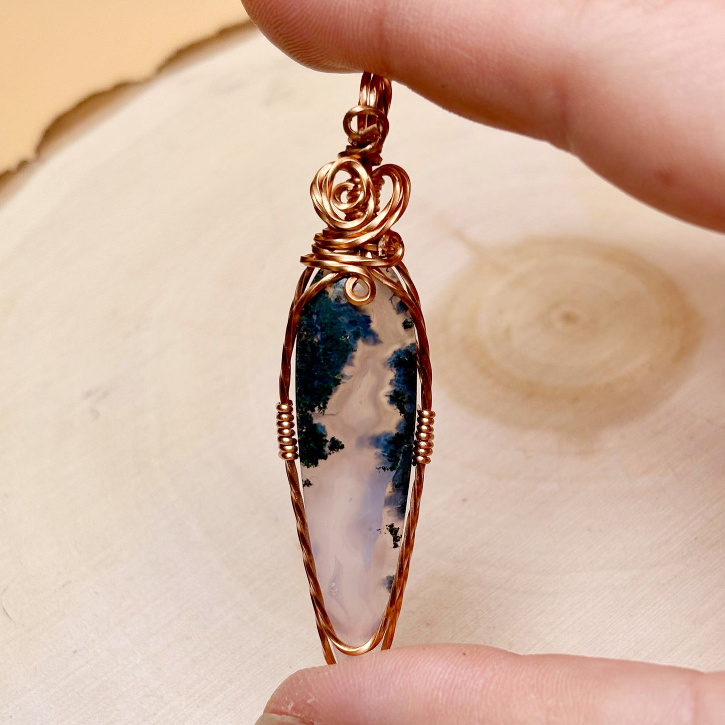 Hand holding a copper-wrapped pendant with a natural stone design on a wooden surface