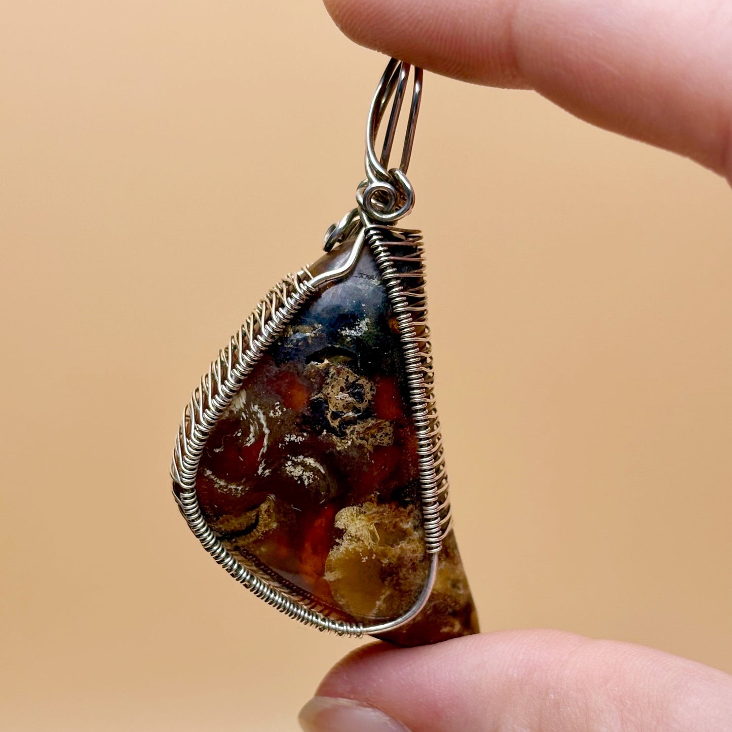 Hand holding a teardrop-shaped pendant with wire wrapping against a beige background