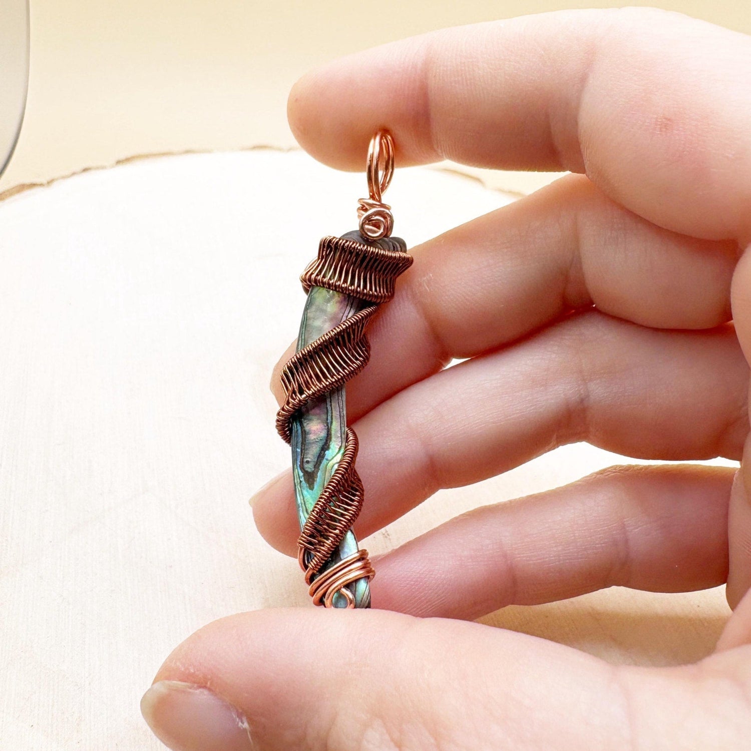 Hand holding a copper-wrapped pendant with a natural shell against a neutral background