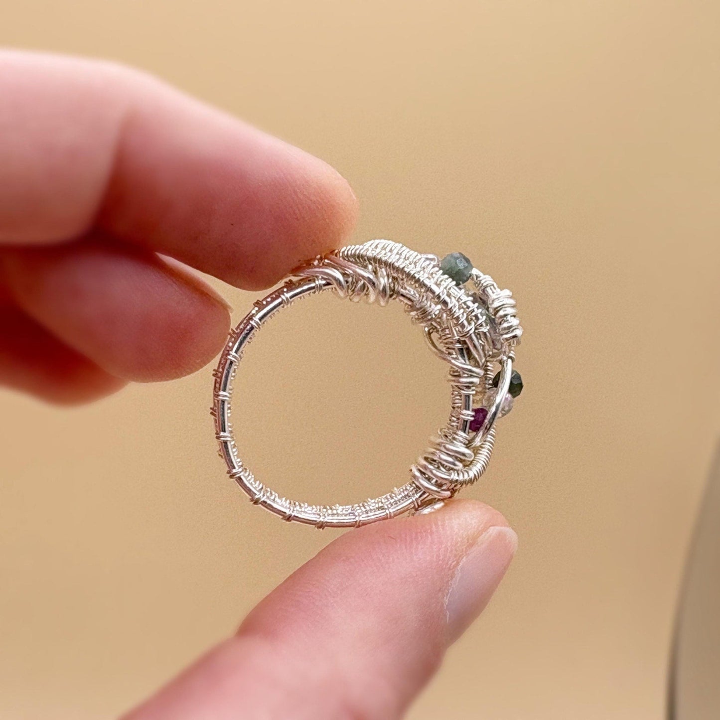 Silver ring with gemstones held between fingers against a beige background
