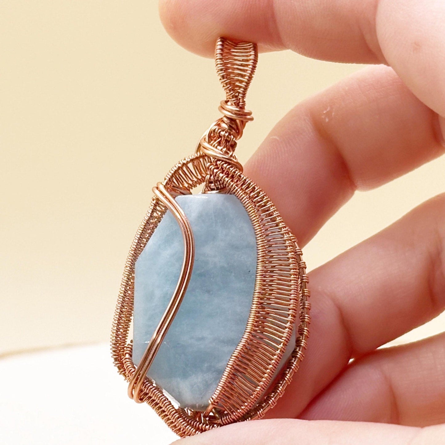 Hand holding a copper-wrapped pendant with a blue stone against a beige background