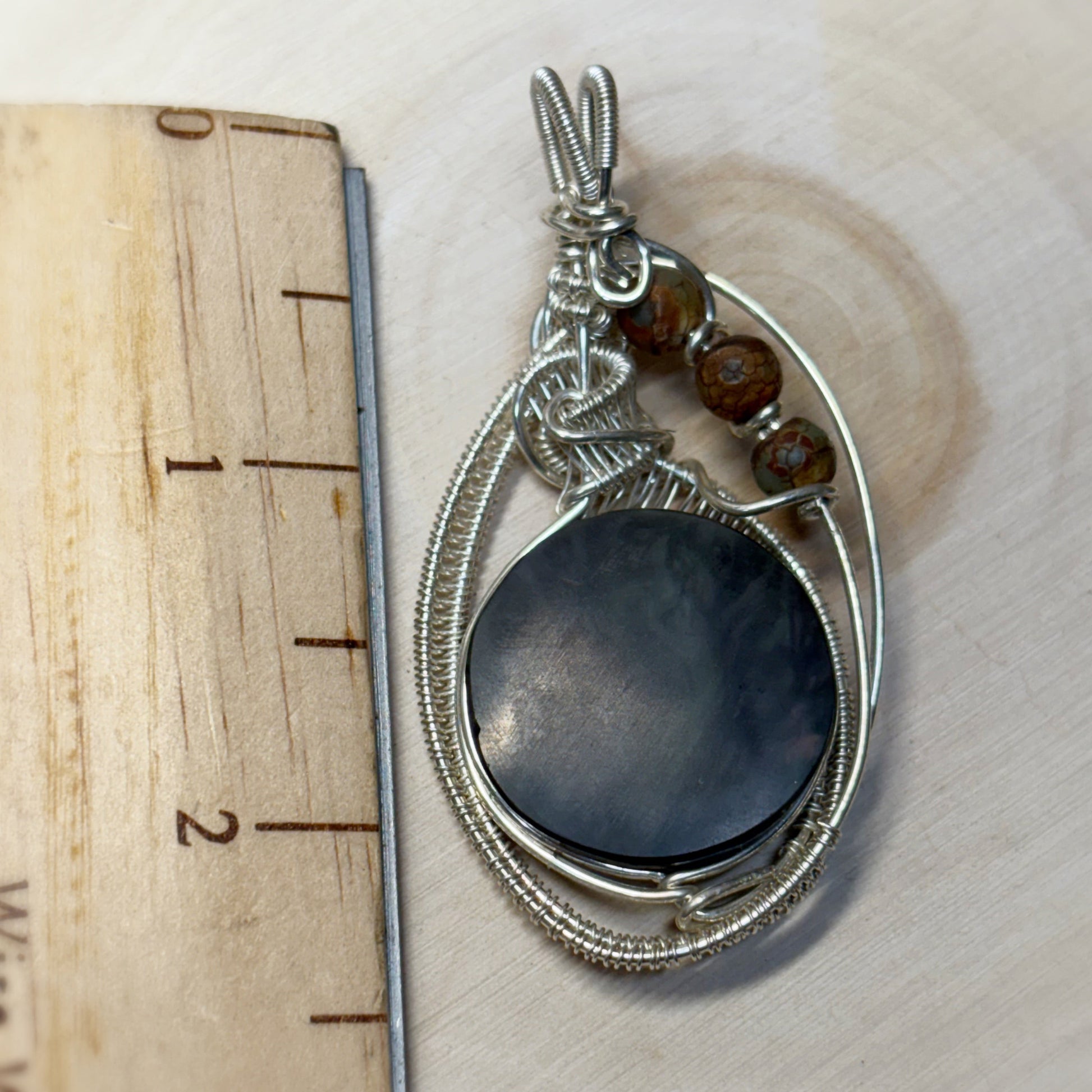 Silver pendant with a dark stone and beads on a wooden surface with a ruler for scale.