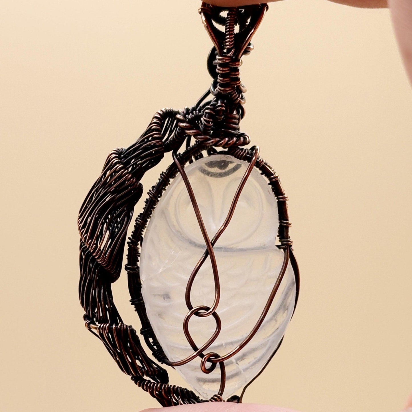 Jewelry pendant with wire wrapping and a central carved owl quartz stone on a beige background