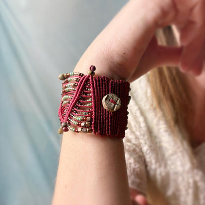 Close-up of a wrist wearing a red macramé bracelet with beads and a button, against a blurred background.