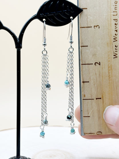 Silver chain earrings with blue beads on a black stand next to a wooden ruler.