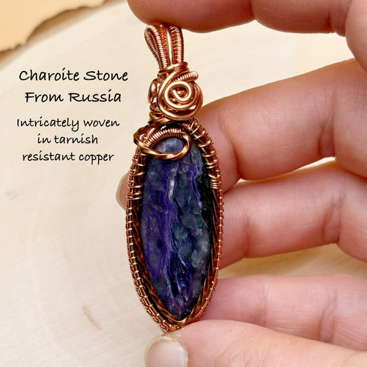 Hand holding a charoite stone pendant with copper wire wrapping, set against a neutral background.