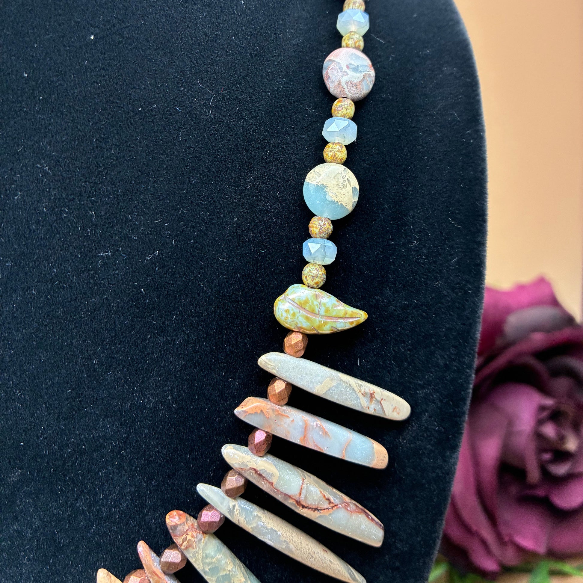 A beaded statement necklace displayed on a black velvet mannequin bust. The necklace features a mix of round, flat, and teardrop-shaped beads in earthy tones of blue, pink, yellow, and brown, with a prominent cluster of elongated stone beads at the bottom.