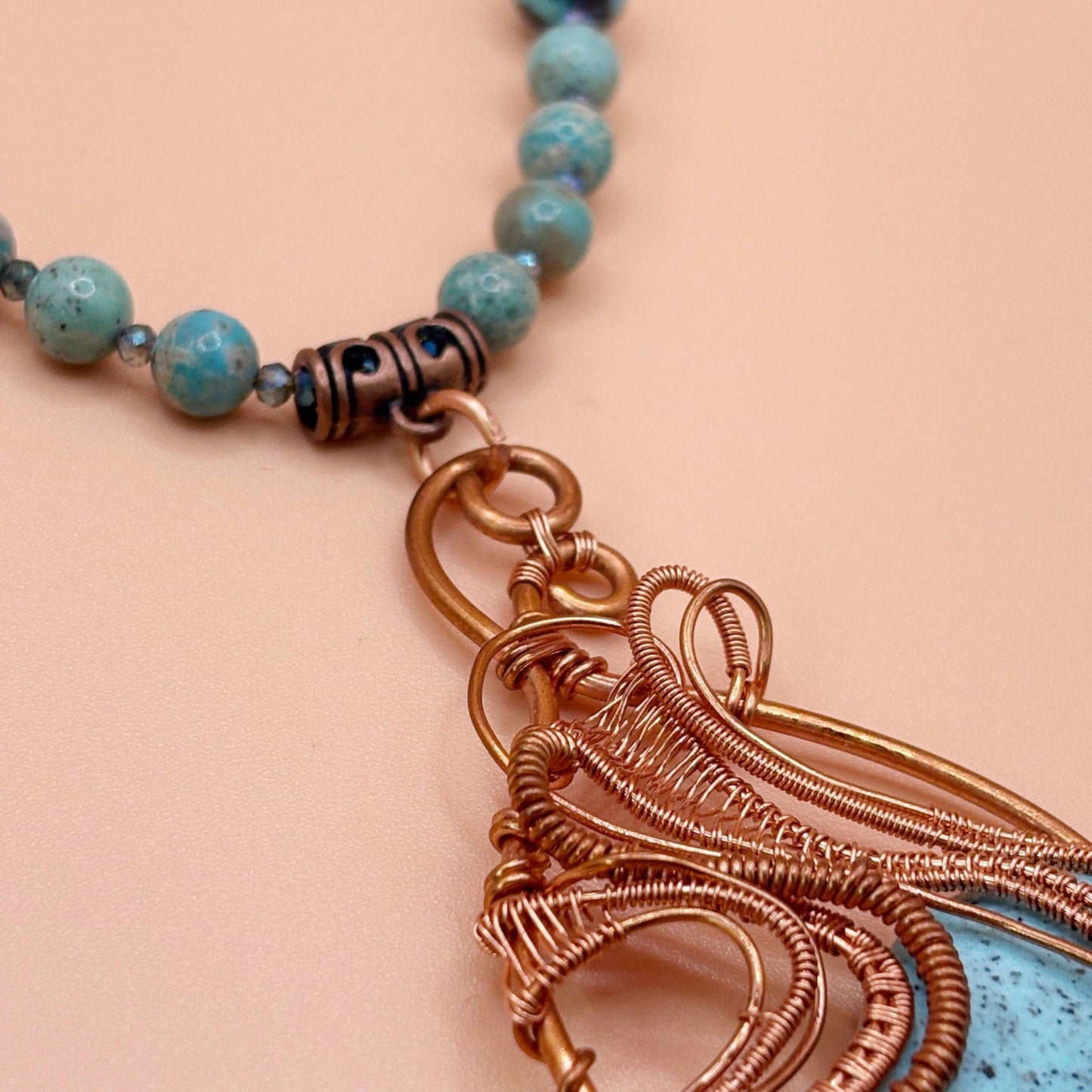 Close-up of a copper wire-wrapped pendant on a turquoise beaded necklace against a pink background