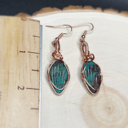 Pair of earrings with green stone and copper wire wrapping on a wooden surface.
