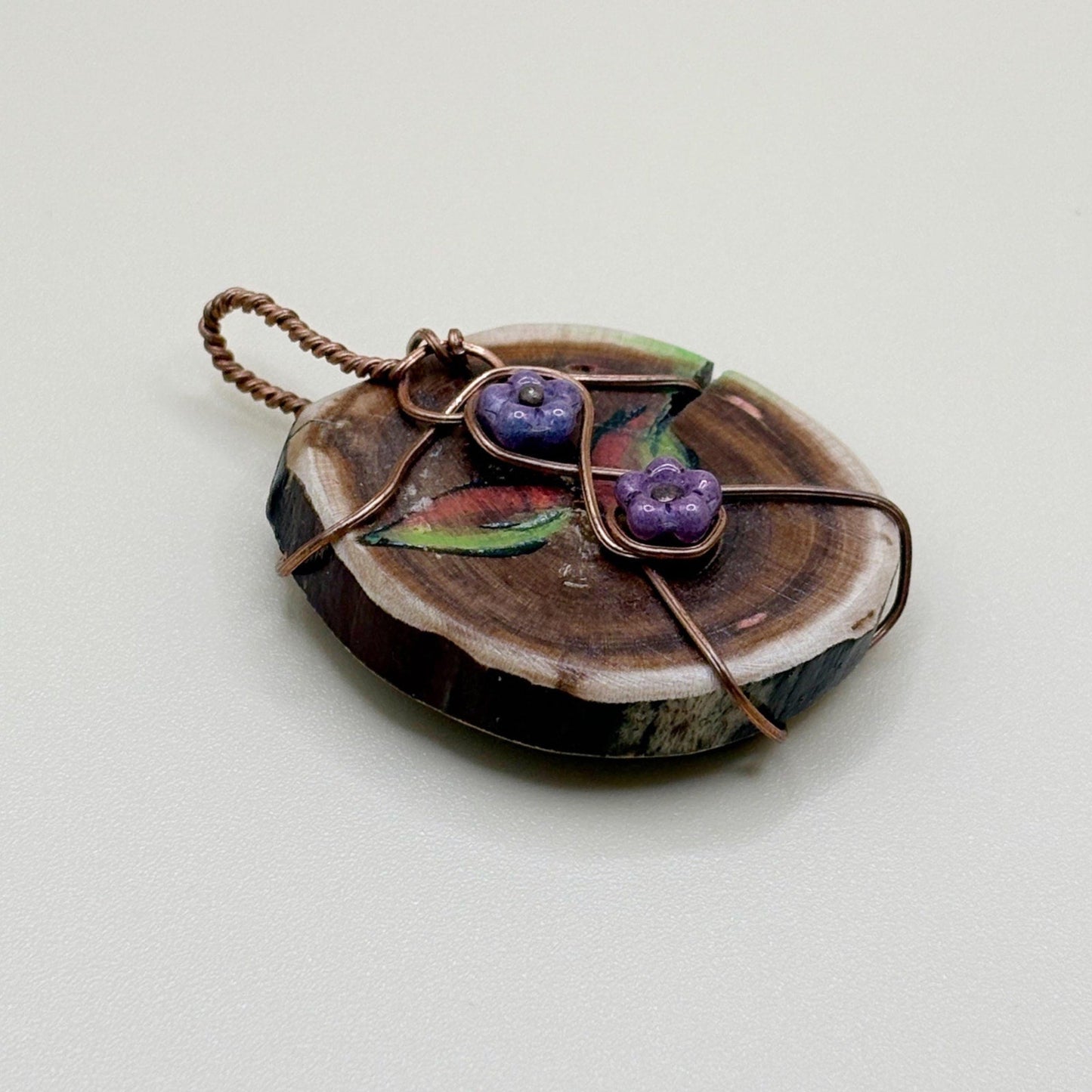 Handmade pendant with purple flowers and copper wire on a wooden slice with a beige background