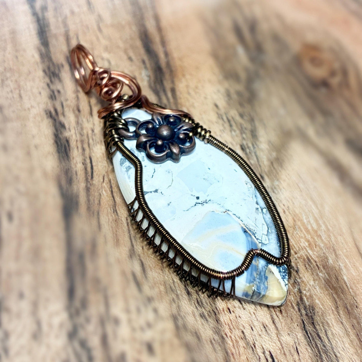 A handmade wire-wrapped pendant featuring a light blue and white marbled stone, accented with dark blue floral details and copper-toned metal, displayed on a rustic wooden surface.