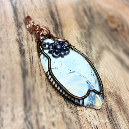 A handmade wire-wrapped pendant featuring a light blue and white marbled stone, accented with dark blue floral details and copper-toned metal, displayed on a rustic wooden surface.