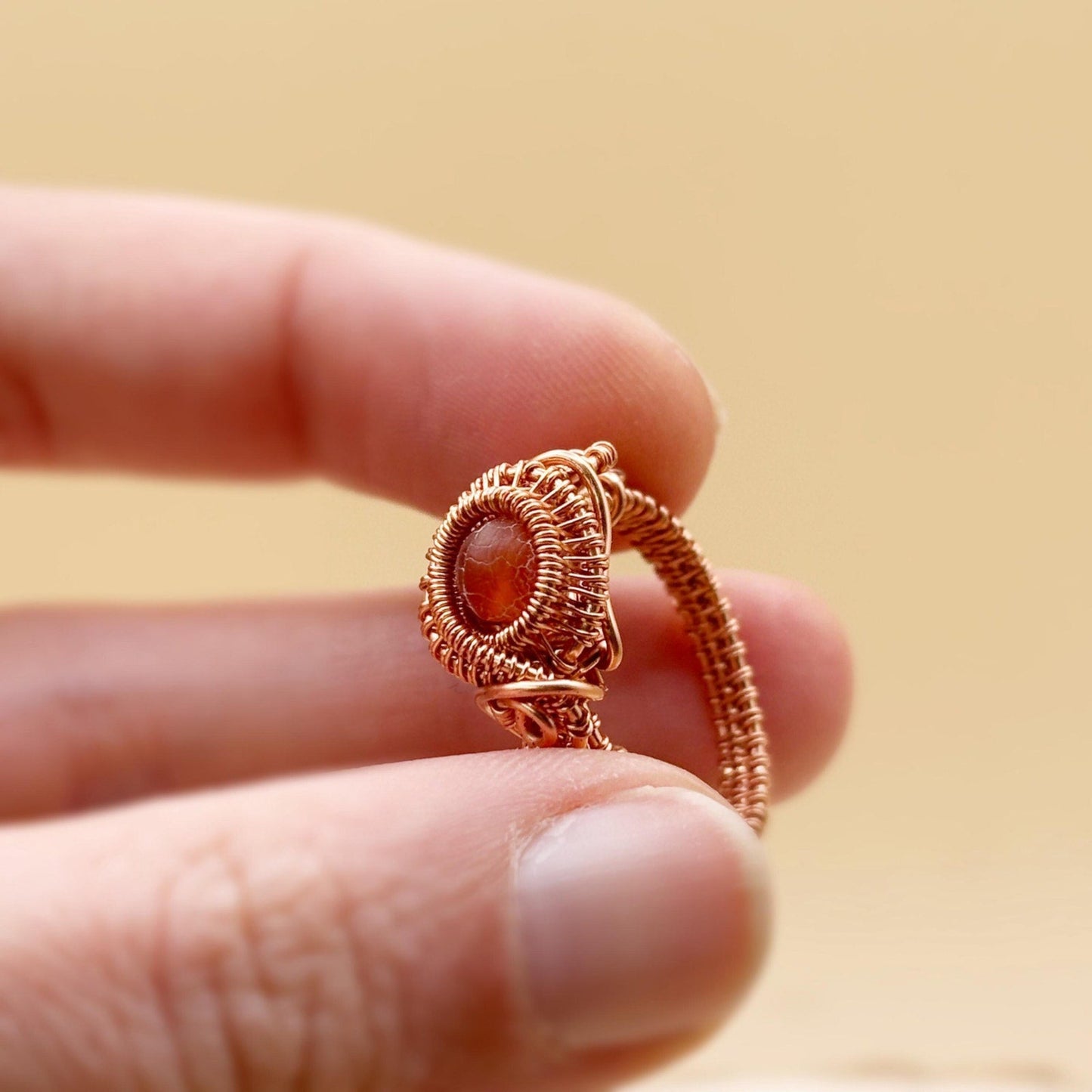 copper wire-wrapped ring with a central gemstone held between fingers against a beige background