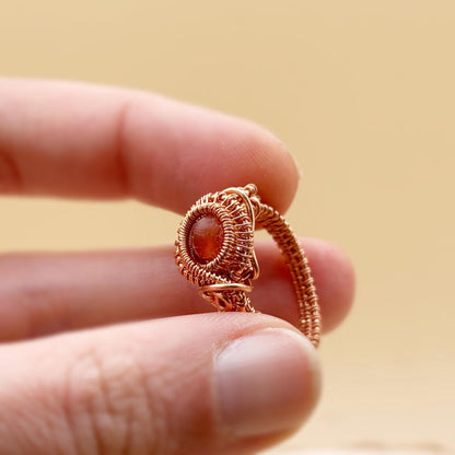 copper wire-wrapped ring with a central gemstone held between fingers against a beige background