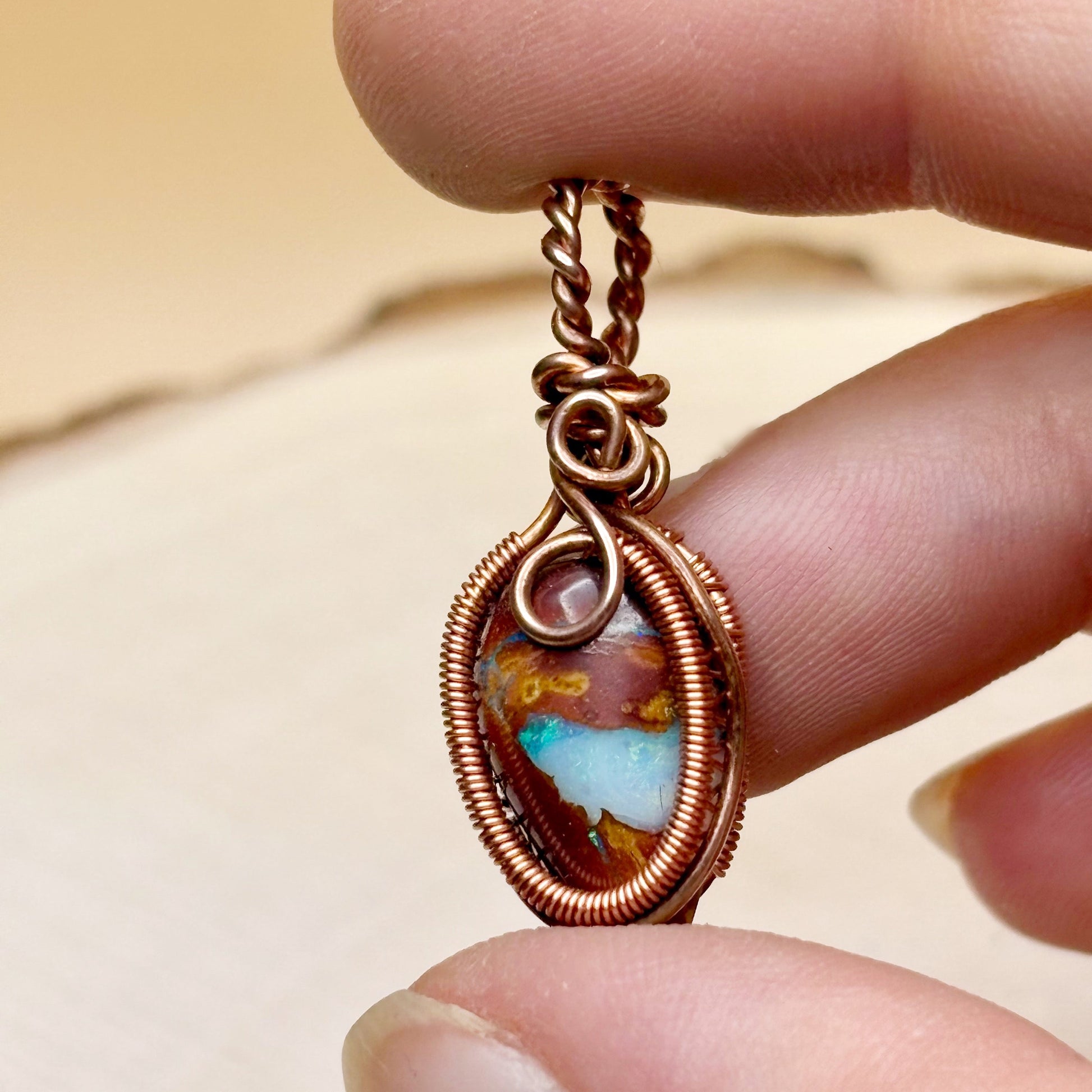 Copper-wrapped pendant with a colorful stone held between fingers against a neutral background