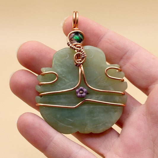 Hand holding a jade pendant with copper wire and gemstones against a beige background