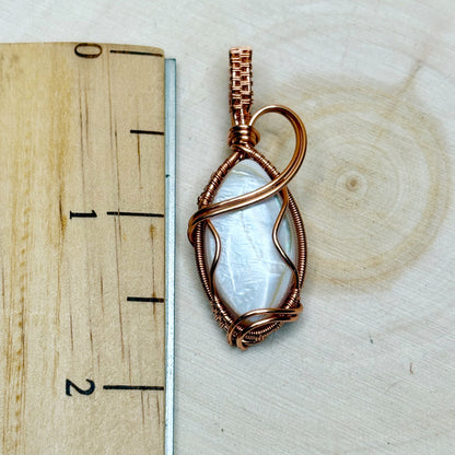 Copper-wrapped pendant with a central mother of pearl shell on a wooden surface with a ruler for scale.