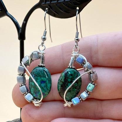 Green stone earrings with silver wire wrapping held in a hand against a beige background
