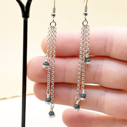 Silver chain earrings with green beads held between fingers against a neutral background