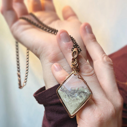 Hand holding a necklace with a square pendant featuring a natural landscape.