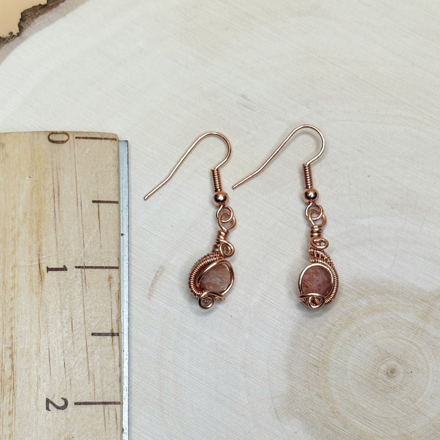 Pair of copper wire-wrapped earrings on a wooden surface with a ruler for scale.