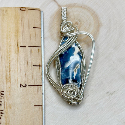 Silver wire-wrapped pendant with a blue and white stone on a wooden surface with a ruler.