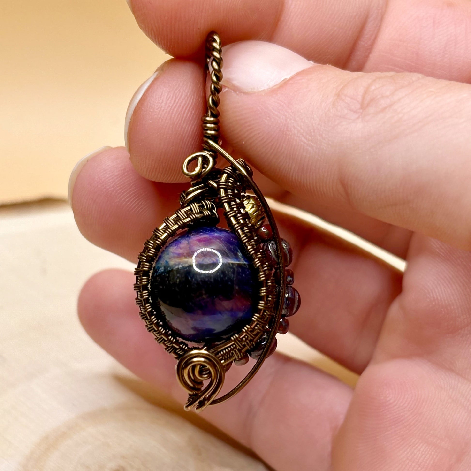 Hand holding a decorative pendant with a stone bead and metal wire wrapping on a wooden surface.