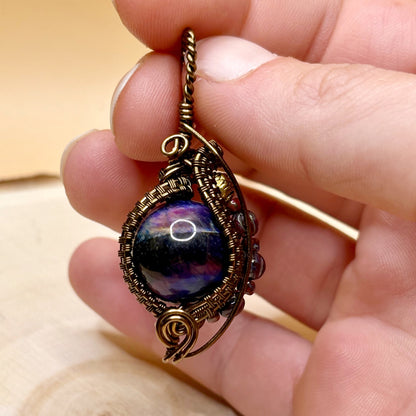 Hand holding a decorative pendant with a stone bead and metal wire wrapping on a wooden surface.