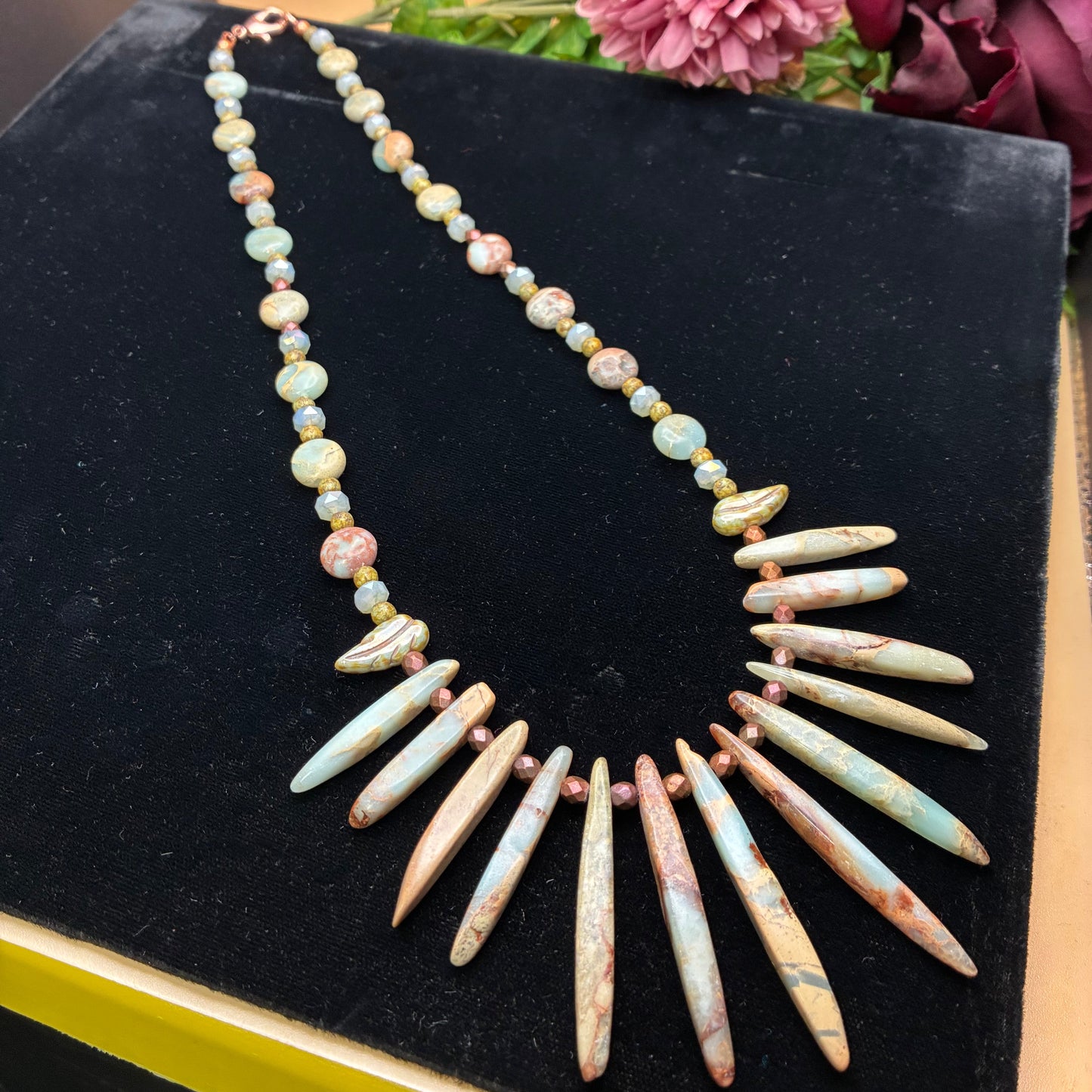 A statement necklace featuring a row of large, pale blue and pink marbled tooth-shaped beads radiating from a beaded chain, displayed on a black velvet jewelry box with flowers in the background.