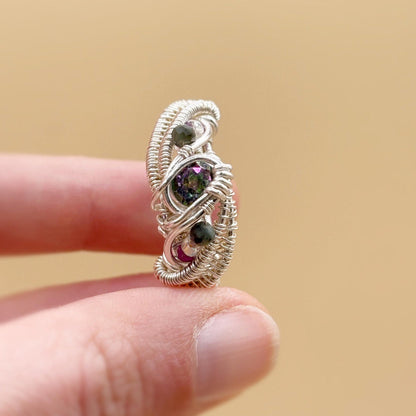 Silver wire-wrapped ring with gemstones held on a finger against a beige background
