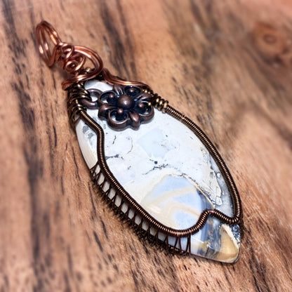 A handmade wire-wrapped pendant featuring a polished white and blue stone with copper-toned metal wrapping, displayed on a rustic wooden surface.