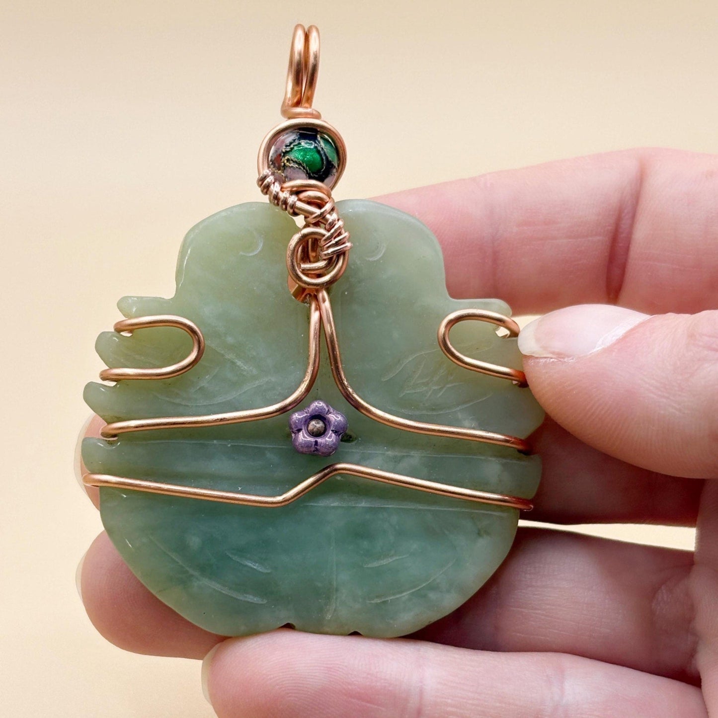 Hand holding a green jade pendant with copper wire wrapping and a small purple flower charm against a beige background.