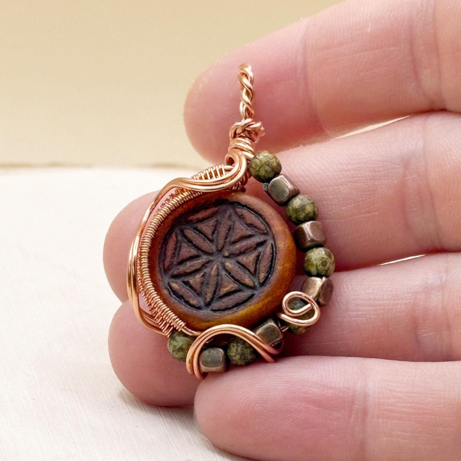 Hand holding a copper pendant with green beads against a neutral background