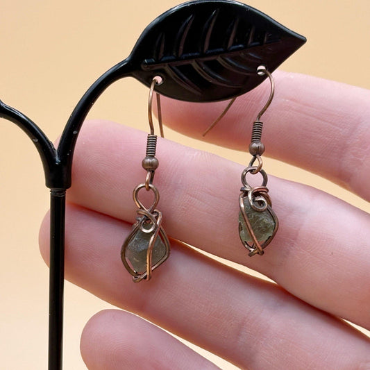 Pair of leaf-shaped moldavite earrings held by a hand against a beige background-p